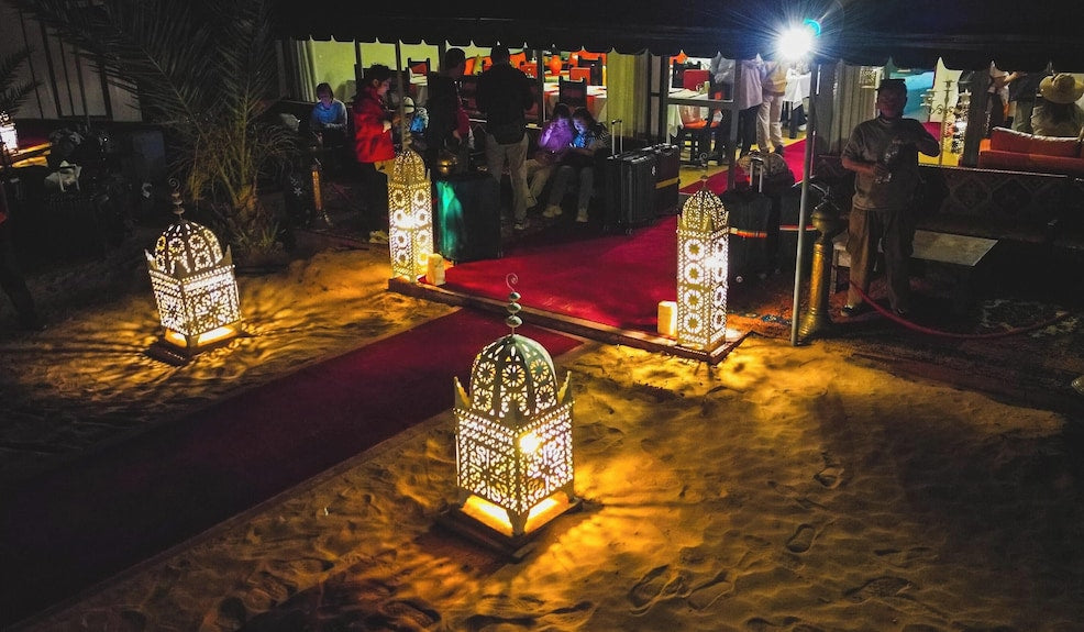 Merzouga Desert Camp night scene with glowing ornate lanterns on sandy ground and people gathered under tents