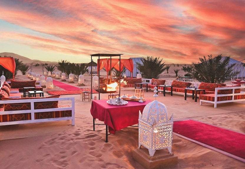 Merzouga Desert Camp outdoor lounge area with red cushions, traditional lanterns, rugs, and desert sunset sky