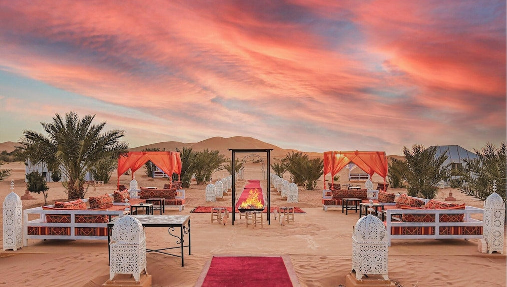 Merzouga Desert Camp outdoor seating area with traditional lanterns, red canopies, patterned cushions, and a central fire pit at sunset