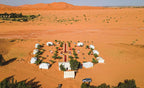 Merzouga Desert Camp with multiple white tents arranged in a circular pattern on orange desert sand surrounded by palm trees under clear sky