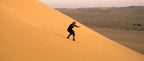 Person wearing black outfit and hiking boots sandboarding down a large golden sand dune in a desert landscape during Merzouga desert tour