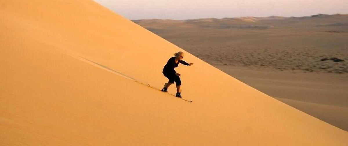 Person wearing black outfit and hiking boots sandboarding down a large golden sand dune in a desert landscape during Merzouga desert tour