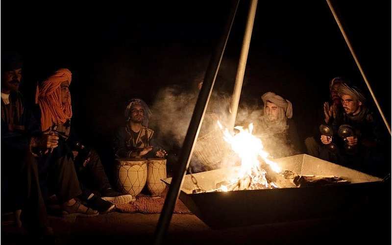 Merzouga Dinner showing a group of men in traditional Berber clothing sitting around a desert campfire playing drums and castanets at night