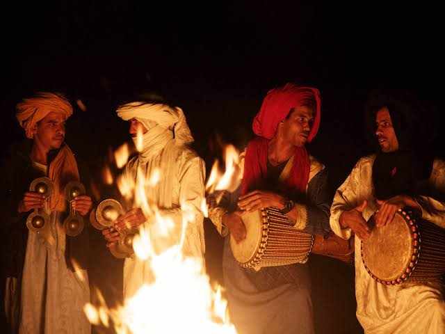 Musicians in traditional Berber attire playing drums and metal castanets around a campfire at night during Merzouga Dinner