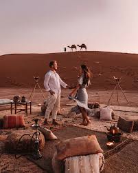 Merzouga Dinner setup with traditional Berber rugs, cushions, low tables, and Moroccan lanterns on desert sand under soft twilight sky