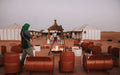 Desert camp seating area with rust-colored barrel chairs, central fire pit, and white Berber-style tents at dusk for Merzouga Dinner.