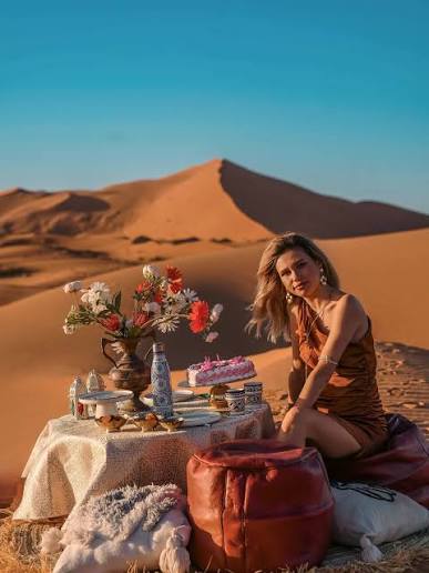Merzouga Dinner setup with a woman sitting by a low table covered in white cloth, decorated with flowers, food, and drink on sand dunes