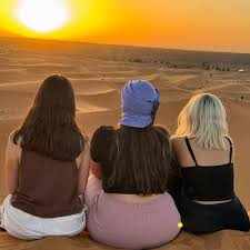 Merzouga Dinner view showing three women sitting on desert sand dunes watching a golden sunset in the Sahara desert