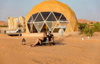 Geodesic yellow dome tent with large windows in the desert, featuring a couple seated on a rug outside near desert plants in a Merzouga Glamping Camp.