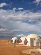 Merzouga Glamping Camp with white dome tents featuring transparent hexagonal windows and doors, set on sandy desert ground under a partly cloudy sky.