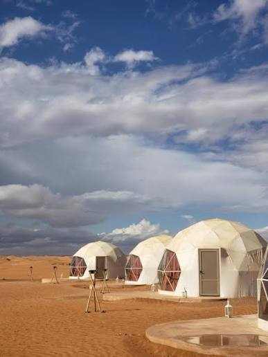 Merzouga Glamping Camp with white dome tents featuring transparent hexagonal windows and doors, set on sandy desert ground under a partly cloudy sky.