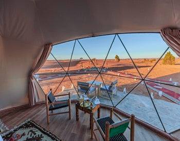 Merzouga Glamping Camp interior with wooden chairs, small table, patterned rug, and panoramic desert view through large dome window.