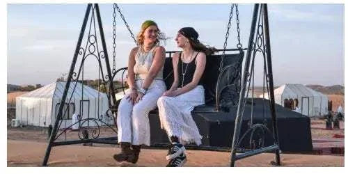 Two women sitting and smiling on a black metal swing with white tents in the background at Merzouga Glamping Camp.