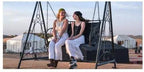 Two women sitting and smiling on a black metal swing with white tents in the background at Merzouga Glamping Camp.