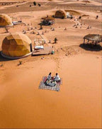 Merzouga Glamping Camp with beige geodesic dome tents and a couple seated on a picnic blanket in the sand desert.