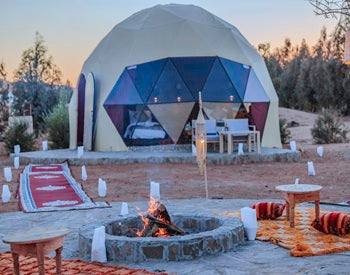Merzouga Glamping Camp featuring a white dome tent with triangular glass windows, outdoor seating, and a central campfire pit.
