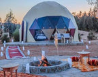 Merzouga Glamping Camp featuring a white dome tent with triangular glass windows, outdoor seating, and a central campfire pit.