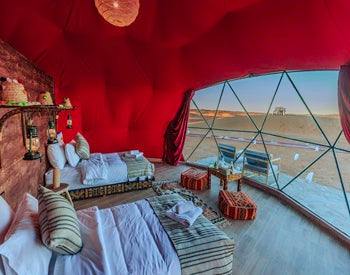 Merzouga Glamping Camp interior featuring spacious red fabric tent with cozy beds, Moroccan textiles, and panoramic desert window view.