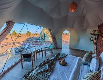 Merzouga Glamping Camp interior featuring a spacious beige tent with large windows, wooden floor, cozy seating, and desert views.