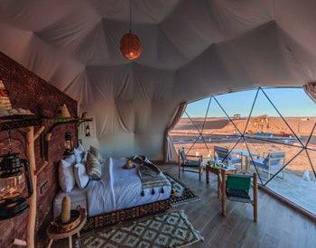 Merzouga Glamping Camp luxury tent interior with white fabric walls, cozy bed, Moroccan rug, wooden furniture, and desert view.