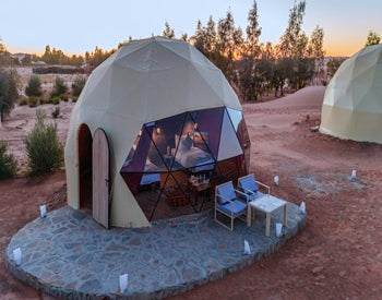 Merzouga Glamping Camp featuring beige dome-shaped tents with wooden doors, outdoor seating, and desert surroundings at sunset.