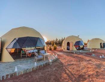 Merzouga Glamping Camp featuring beige dome tents with geometric dark window panels, outdoor seating, set on reddish desert ground.