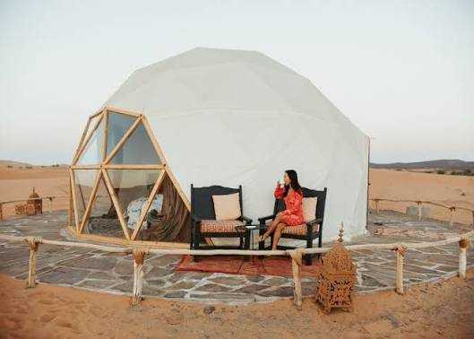 Geodesic dome tent with white fabric and wooden frame, outdoor seating, and a woman relaxing at a Merzouga Glamping Camp in the desert.