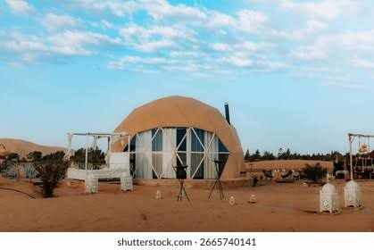 Merzouga Glamping Camp dome tent in beige with glass panels, situated in a sandy desert with lantern decorations and seating area nearby.