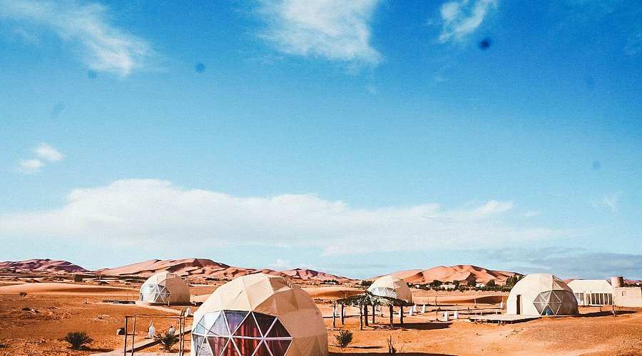 Merzouga Glamping Camp featuring white geodesic dome tents with transparent triangular windows set in sandy desert dunes.