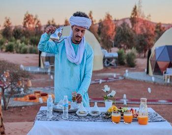Merzouga Glamping Camp desert staff in blue traditional attire preparing tea on a table with glasses and refreshments outdoors at sunset.
