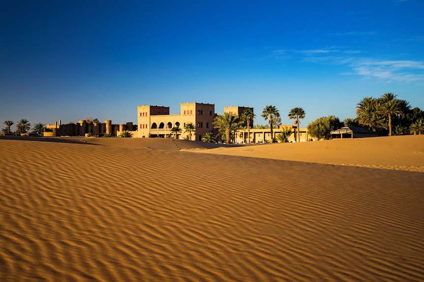 Merzouga hotel kanz Erremal sandy desert landscape with traditional Moroccan architecture and palm trees under a clear sky