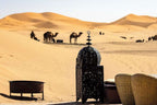 Merzouga hotel kanz Erremal outdoor desert seating area with woven chairs, metal lantern, and camels in the sand dunes