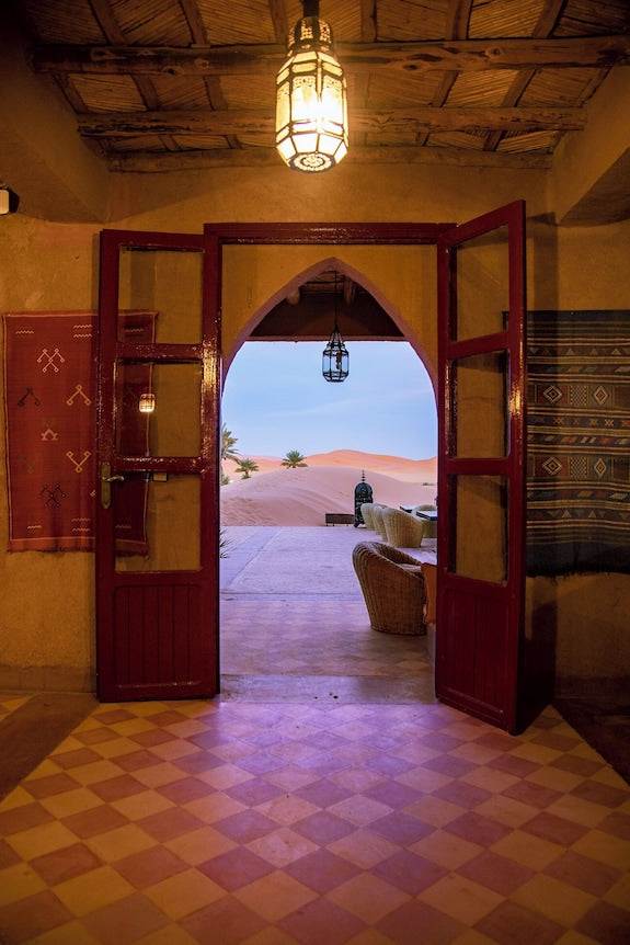 Merzouga hotel kanz Erremal entrance with traditional red wooden doors opening to desert sand dunes and lantern lighting