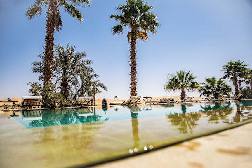 Merzouga hotel kanz Erremal outdoor pool with clear water, surrounded by palm trees and lounge chairs under a bright blue sky