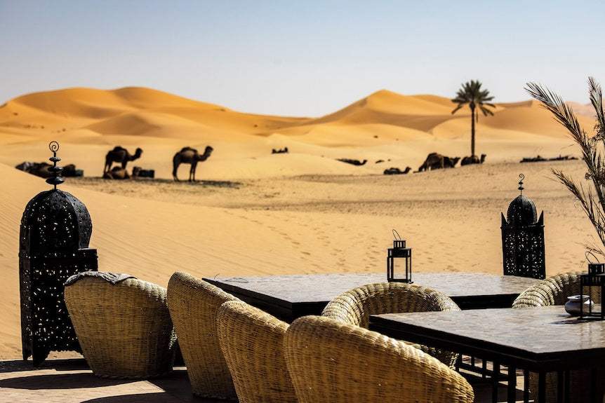 Outdoor dining area with wicker chairs and tables featuring lanterns against desert sand dunes and camels at Merzouga hotel kanz Erremal