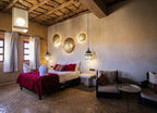 Merzouga hotel kanz Erremal room with rustic beige walls, woven ceiling, dark wood bed with red and white bedding, lantern lights, and cushioned seating