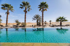 Swimming pool with clear blue water surrounded by palm trees and lounge chairs at Merzouga hotel kanz Erremal desert resort