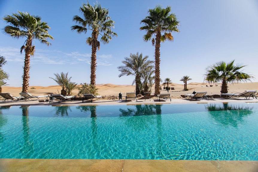 Swimming pool with clear blue water surrounded by palm trees and lounge chairs at Merzouga hotel kanz Erremal desert resort