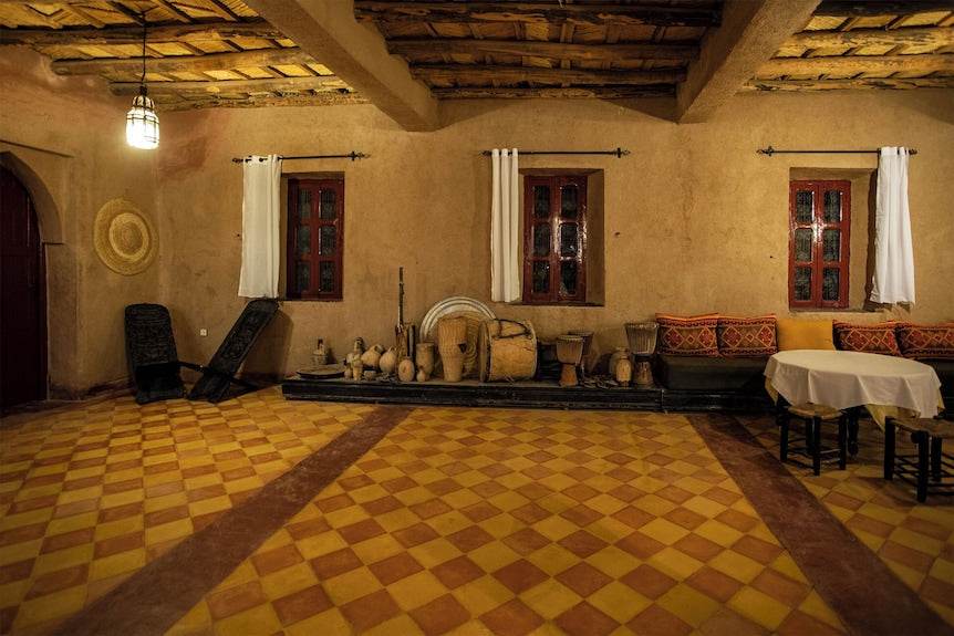 Traditional Moroccan interior in Merzouga hotel kanz Erremal with warm earth tones, clay walls, red windows, rustic ceiling, and patterned tile floor