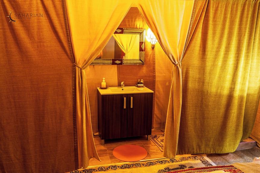 Merzouga Luxury Camp bathroom area with wooden vanity, white sink, mirror with patterned frame, orange rug, and yellow curtains