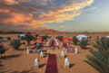 Merzouga Luxury Camp setup in the desert with red carpet, white lanterns, tents, palm trees, and golden sand dunes under a colorful sky