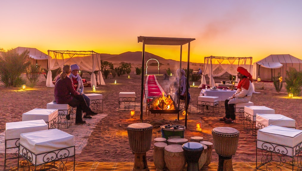 Merzouga Luxury Camp desert seating area with cushioned stools, central fire pit, traditional drums, lit lanterns at sunset