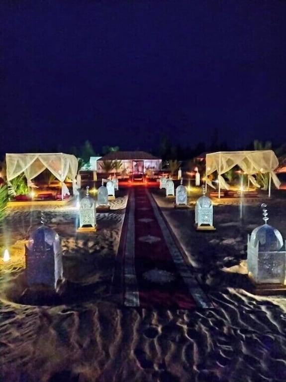 Merzouga Luxury Camp at night showing illuminated lanterns, carpeted walkway, and white canopy tents on desert sand