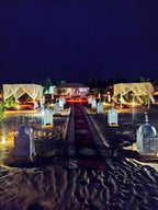 Merzouga Luxury Camp at night showing illuminated lanterns, carpeted walkway, and white canopy tents on desert sand