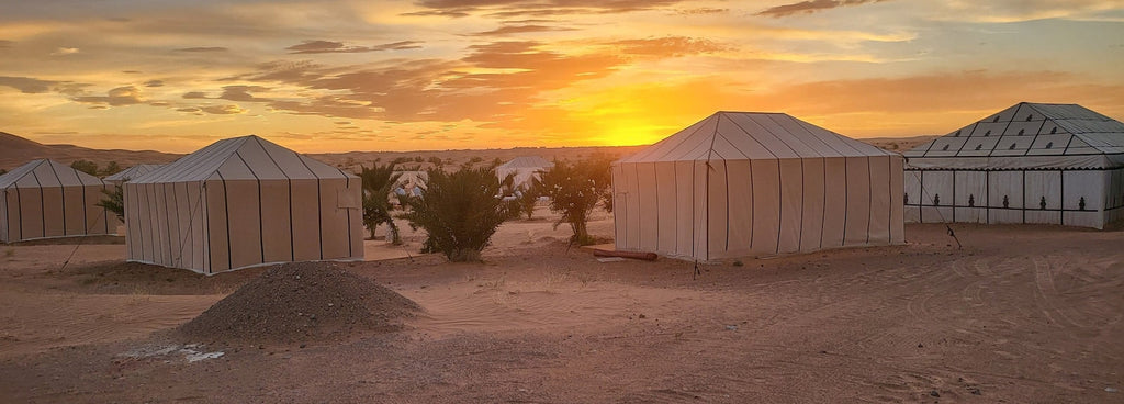 Merzouga Luxury Camp at sunset featuring white desert tents with vertical stripes and a sandy landscape with sparse vegetation