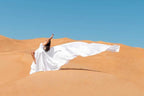 Woman in flowing white dress striking a pose on golden desert dunes under clear blue sky at Merzouga Luxury Camp