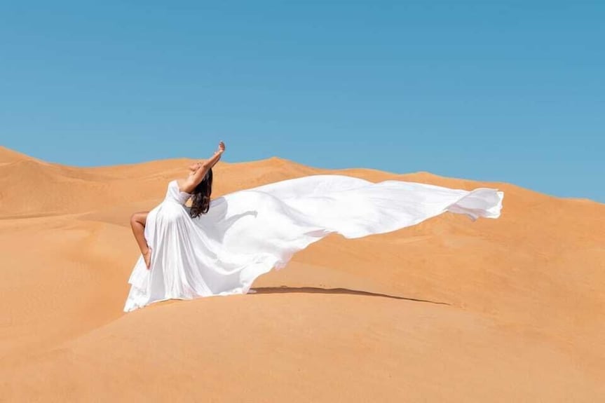 Woman in flowing white dress striking a pose on golden desert dunes under clear blue sky at Merzouga Luxury Camp