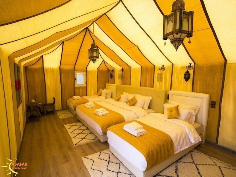 Merzouga Luxury Camp interior with warm yellow canvas tent, three beds, modern decor, wooden flooring and hanging lanterns