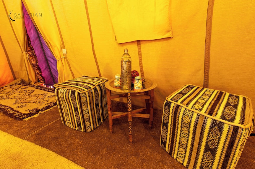 Merzouga Luxury Camp interior with yellow fabric walls, patterned ottomans, small wooden table, and traditional desert decor