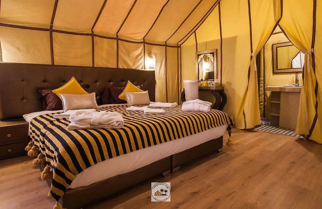 Luxury desert tent interior featuring a large bed with black and yellow striped blanket and tan curtains at Merzouga Overnight Luxury Sahara Desert Camp.